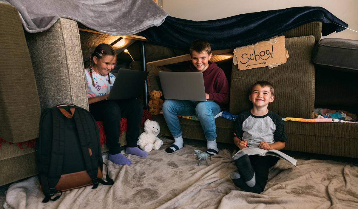 Three elementary-aged students learning remotely in pillow fort at home. 